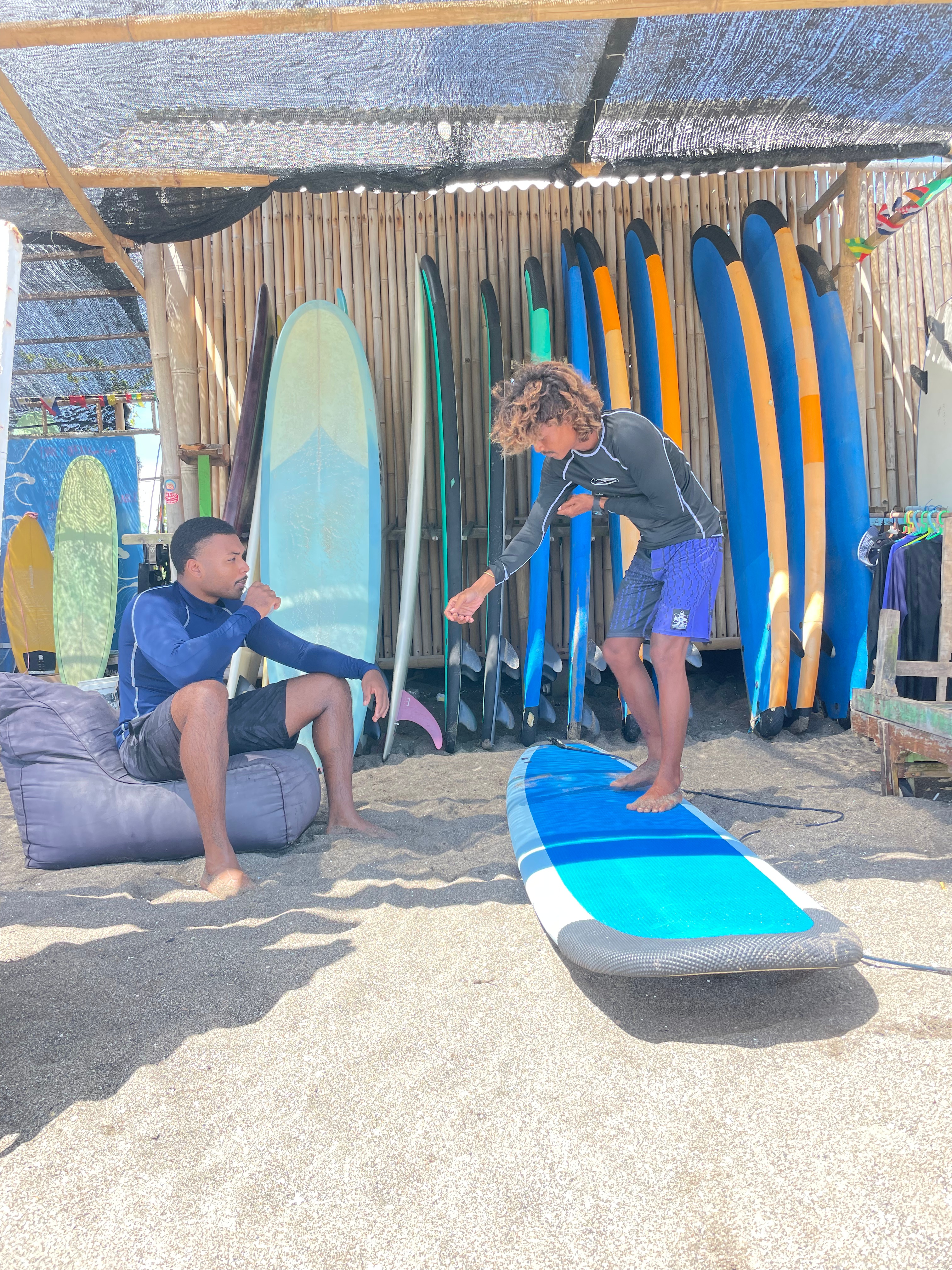 Surf student sitting on a bean bag during a theory lesson, while the instructor stands on a soft-top board demonstrating proper positioning, with surfboards displayed on the wall in the background.