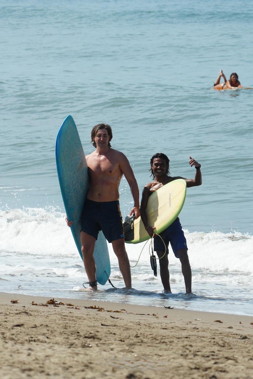 Surf instructor and student smiling for a photo after their surf lesson, holding surfboards with the ocean in the background.